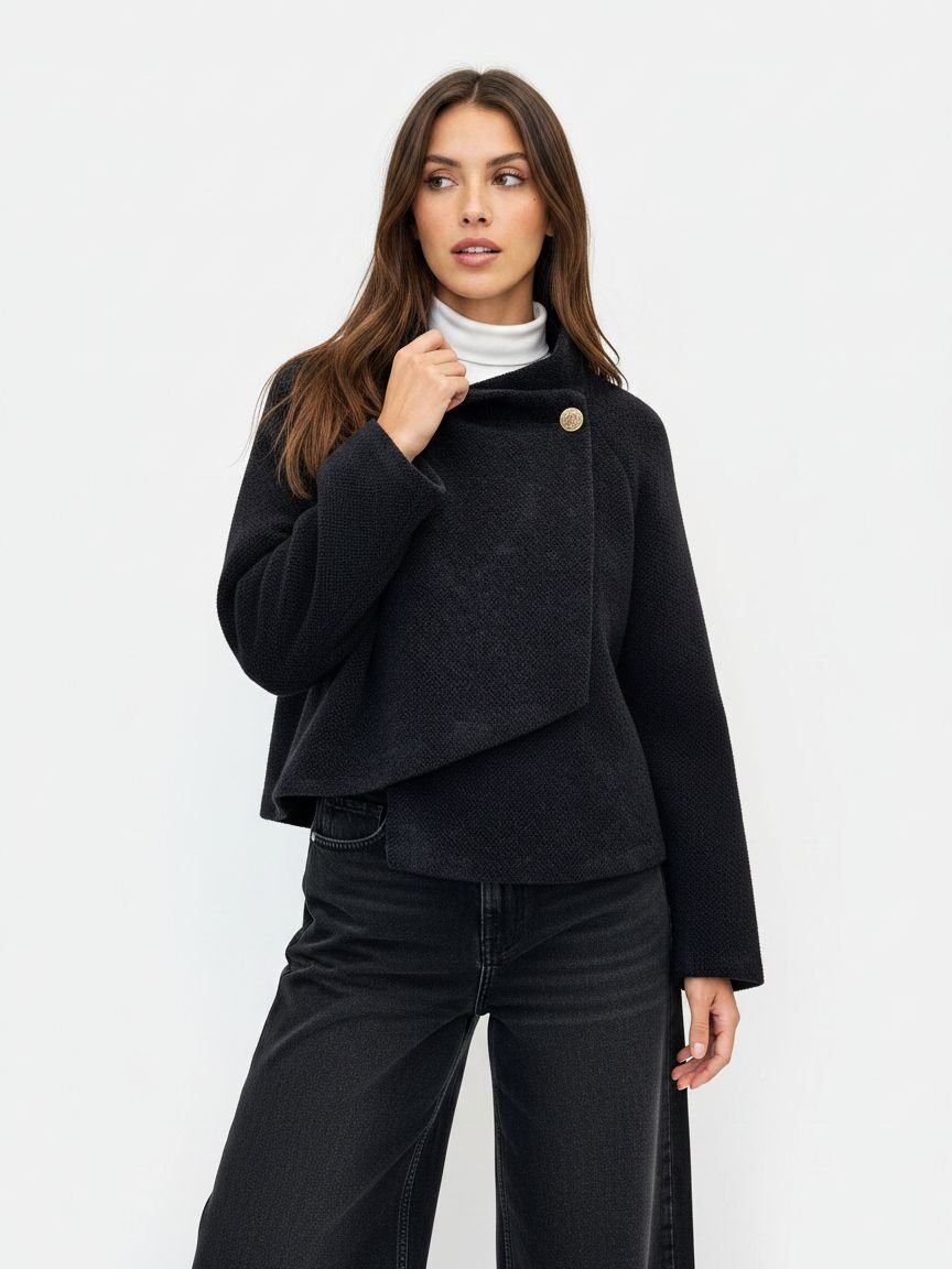 A woman modeling a black asymmetrical wrap-style jacket with a single gold-tone button, worn over a white turtleneck and black wide-leg jeans, against a plain white studio background.