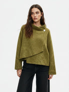 A woman wearing a textured olive green asymmetrical wrap sweater with a single decorative button, paired with black wide-leg jeans, against a plain white studio background.