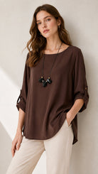 A woman wearing a dark brown, loose-fitting long-sleeve top with rolled cuffs, paired with cream pants and a black pendant necklace, posing against a neutral light beige wall.