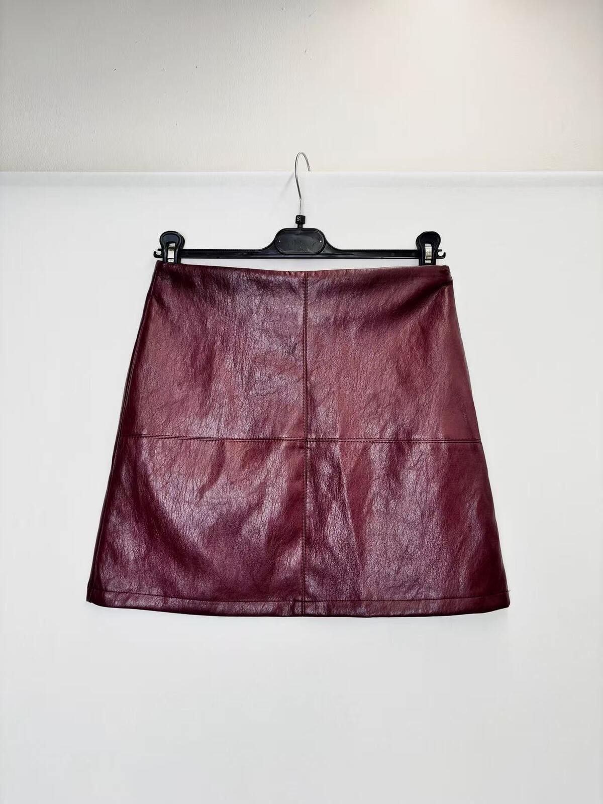 A dark burgundy faux leather A-line mini skirt hanging on a black hanger against a plain white wall.