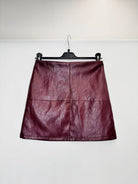 A dark burgundy faux leather A-line mini skirt hanging on a black hanger against a plain white wall.