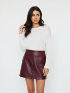 A woman modeling a white long-sleeve knit top and a dark burgundy faux-leather mini skirt against a plain white studio background.