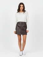 A woman modeling a dark brown faux-leather mini skirt paired with a white long-sleeve top and white sneakers, standing against a plain white studio background.
