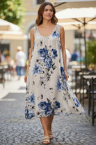 A woman wearing a sleeveless white midi dress with a blue floral print, walking on a cobblestone street in an outdoor cafe setting.