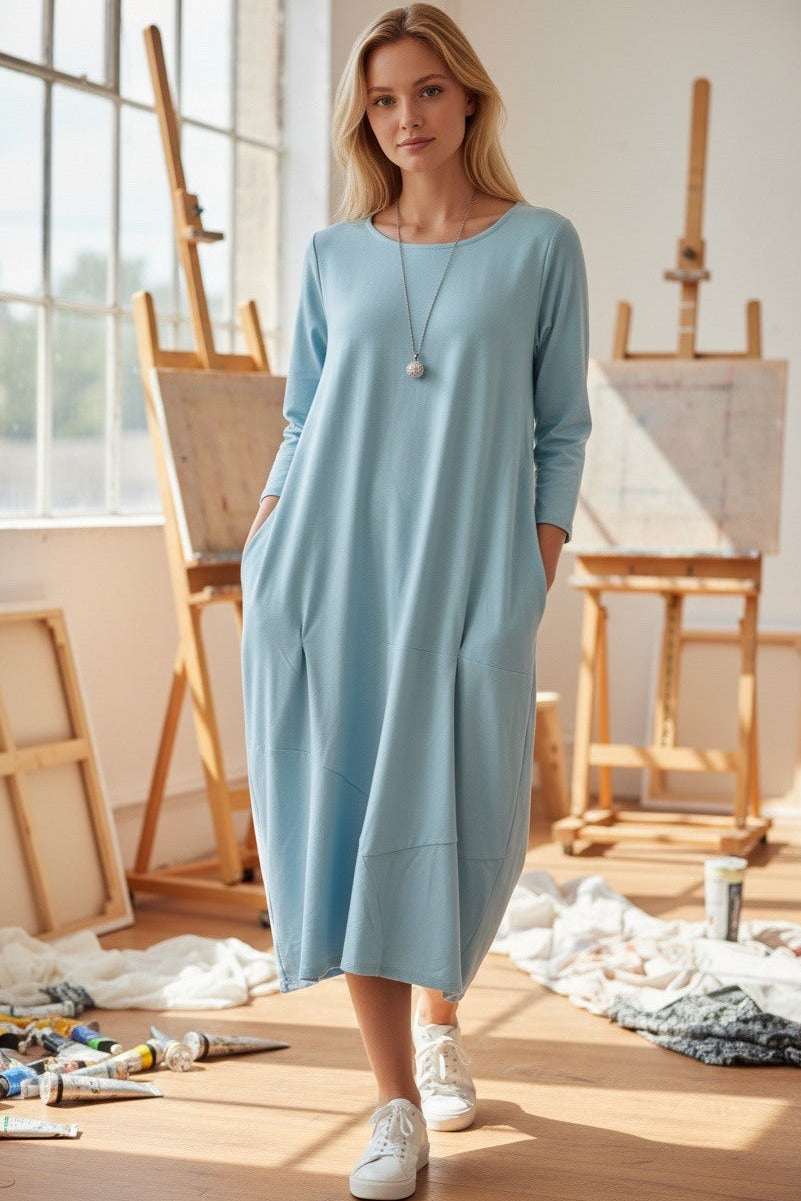 Woman in a light blue dress standing in an artist's studio with easels and art supplies.