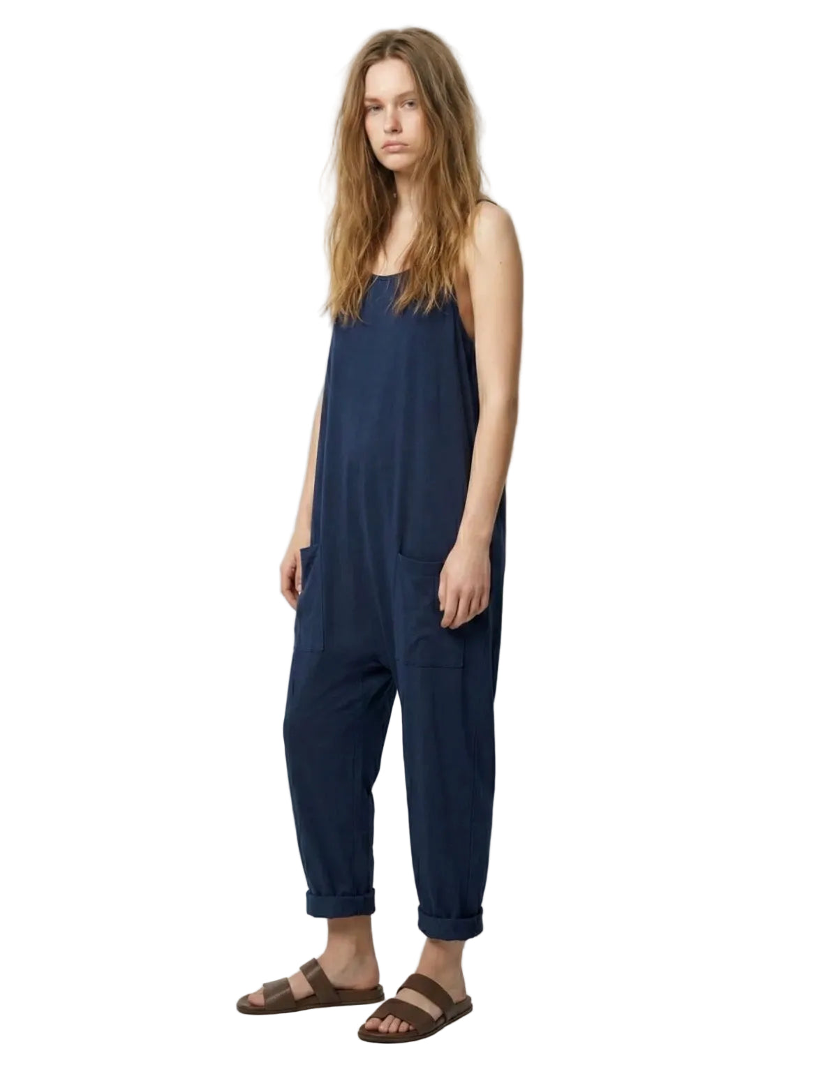 A woman modeling a navy blue sleeveless jumpsuit with large front patch pockets and cuffed ankles, paired with brown slide sandals, against a plain light gray studio background.