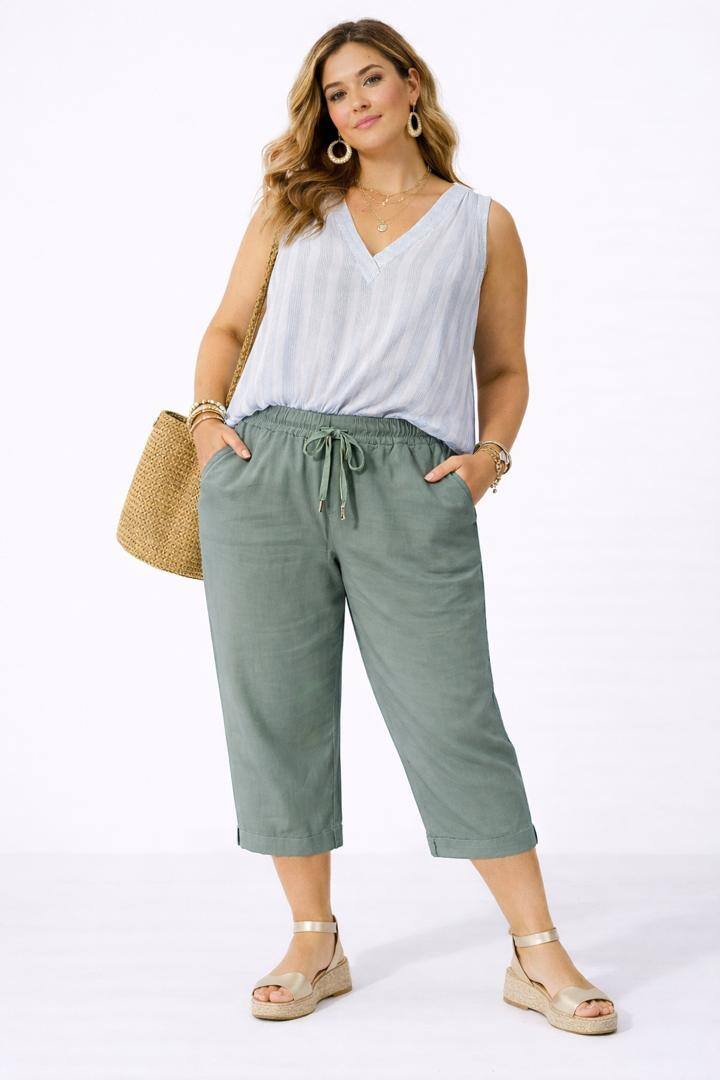 Woman wearing a white sleeveless top and olive green pants with a straw bag on a white background