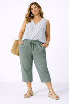 Woman wearing a white sleeveless top and olive green pants with a straw bag on a white background