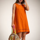 A woman wearing a sleeveless orange linen dress, holding a woven straw tote bag with colorful striped handles, against a plain light background.