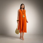 A woman models a sleeveless bright orange midi dress, holding a small woven straw bag. 