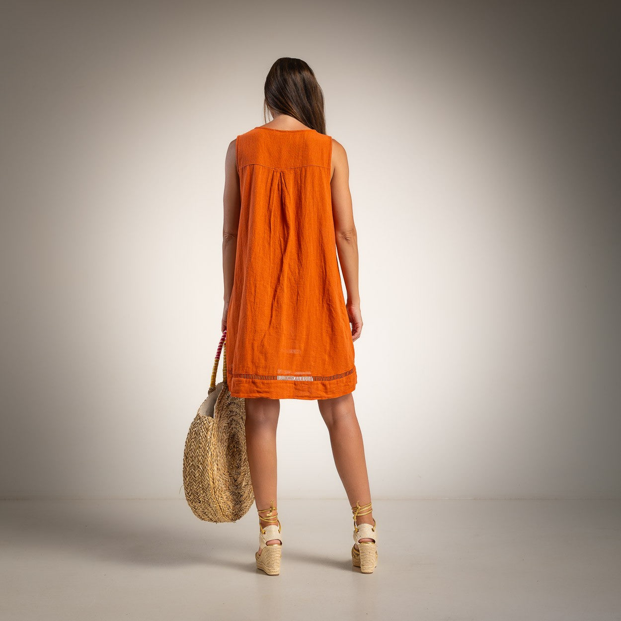 A woman is shown from behind wearing a sleeveless orange dress and holding a woven straw tote bag against a neutral studio background.
