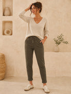 A woman stands in a warm, neutral-toned room wearing a light beige V-neck knit sweater, olive green drawstring pants with textured side panels, and white sneakers.