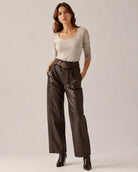 A woman modeling a beige long-sleeve scoop-neck top and dark brown wide-leg faux-leather trousers with a matching belt, paired with black ankle boots