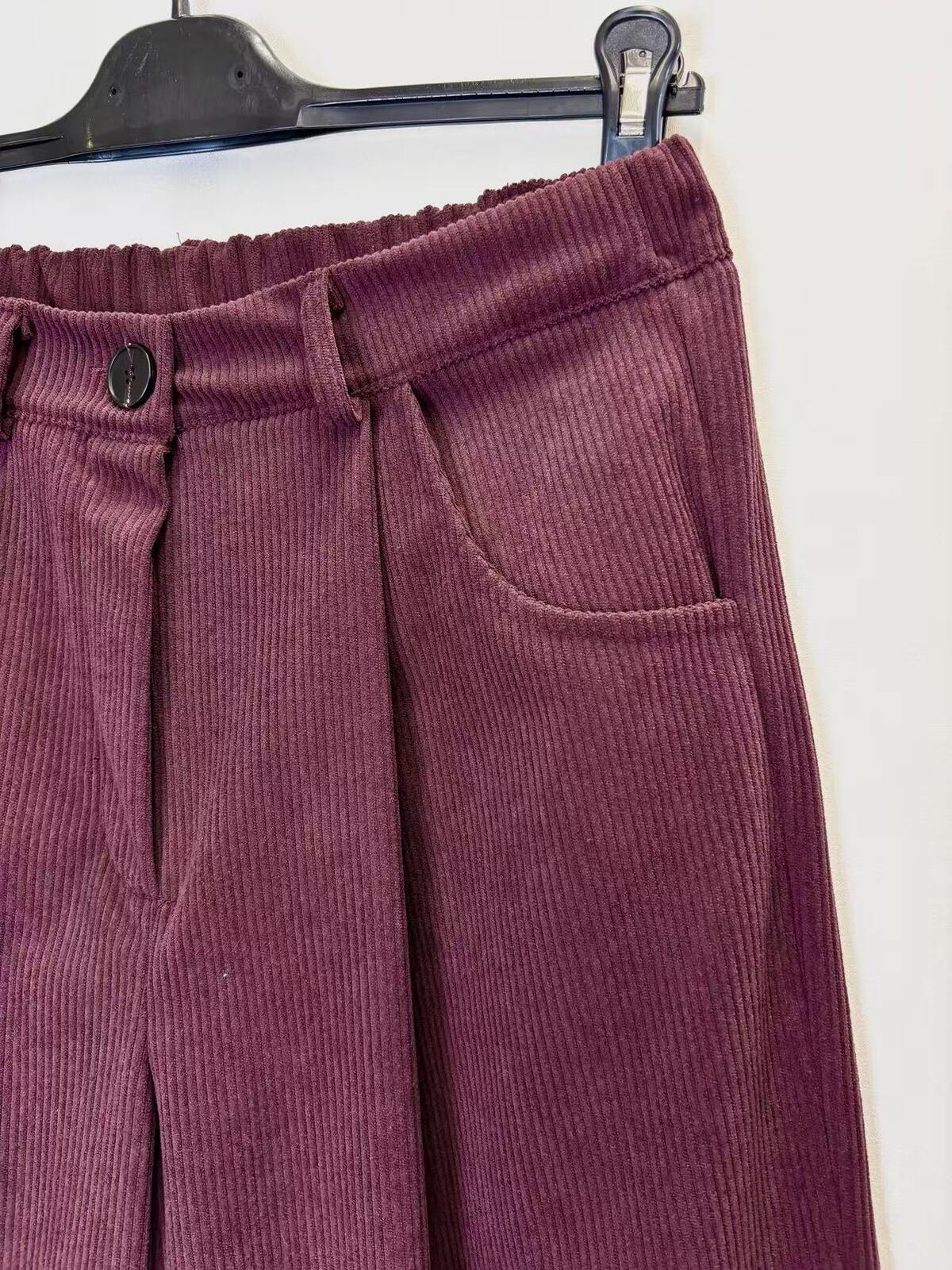 A close-up of a pair of corduroy pants in a deep plum purple color, hanging on a black hanger against a plain white wall.