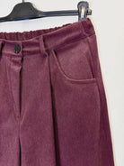 A close-up of a pair of corduroy pants in a deep plum purple color, hanging on a black hanger against a plain white wall.