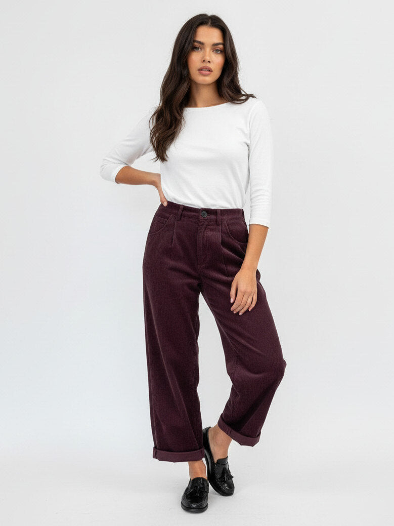 A woman modeling a pair of dark burgundy corduroy wide-leg cropped pants with a white long-sleeve top and black loafers, standing against a plain white studio background.