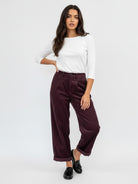 A woman modeling a pair of dark burgundy corduroy wide-leg cropped pants with a white long-sleeve top and black loafers, standing against a plain white studio background.
