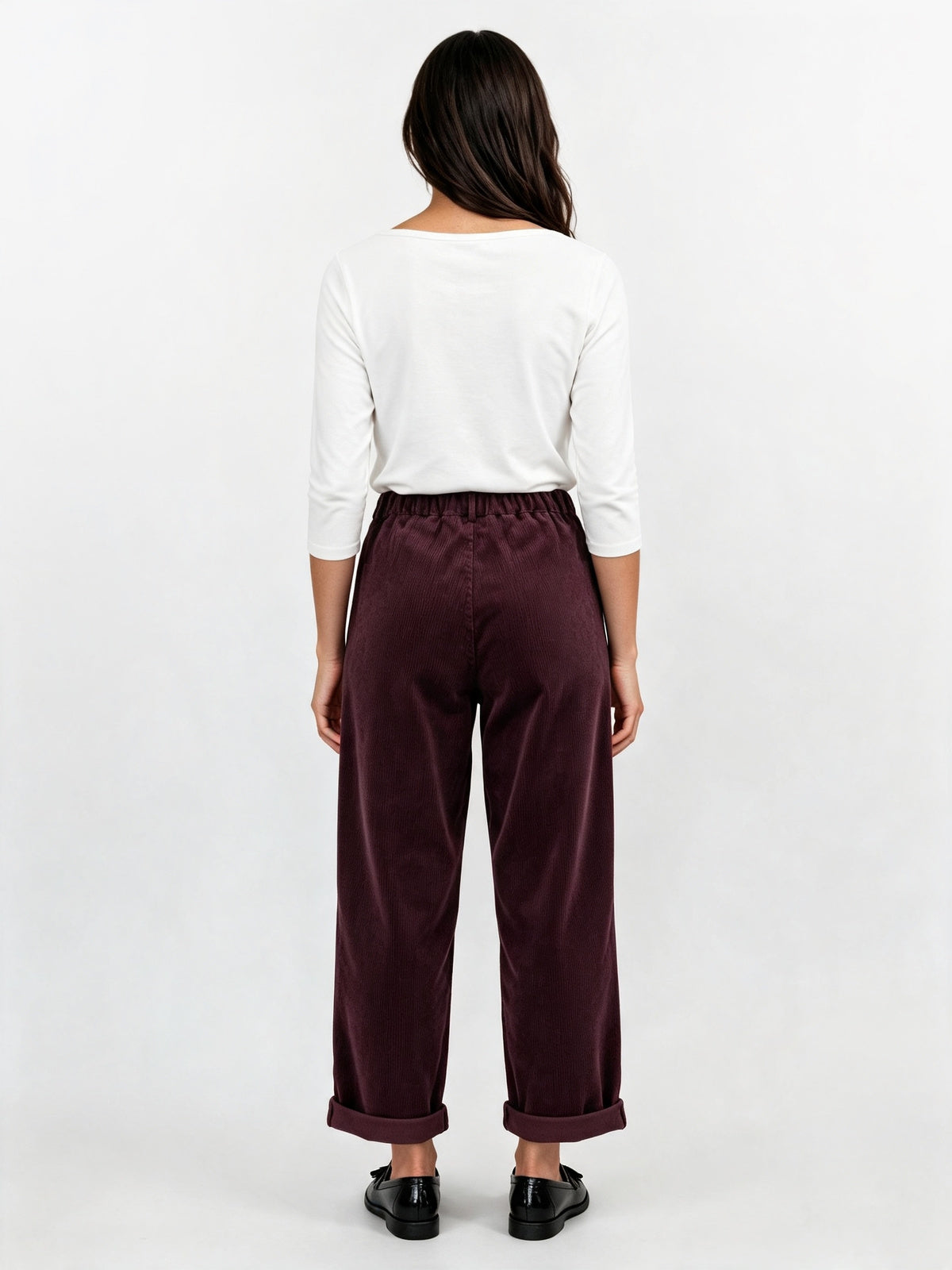 A woman viewed from the back wearing a white long-sleeve top and dark burgundy wide-leg trousers with black shoes, against a plain white studio background.