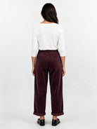 A woman viewed from the back wearing a white long-sleeve top and dark burgundy wide-leg trousers with black shoes, against a plain white studio background.