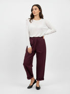 A woman modeling a white long-sleeve top and dark burgundy cropped trousers with black flats, standing against a plain light gray studio background.