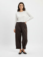 A woman modeling a white long-sleeve top and dark brown wide-leg cropped pants with black flats, standing against a light gray studio background.