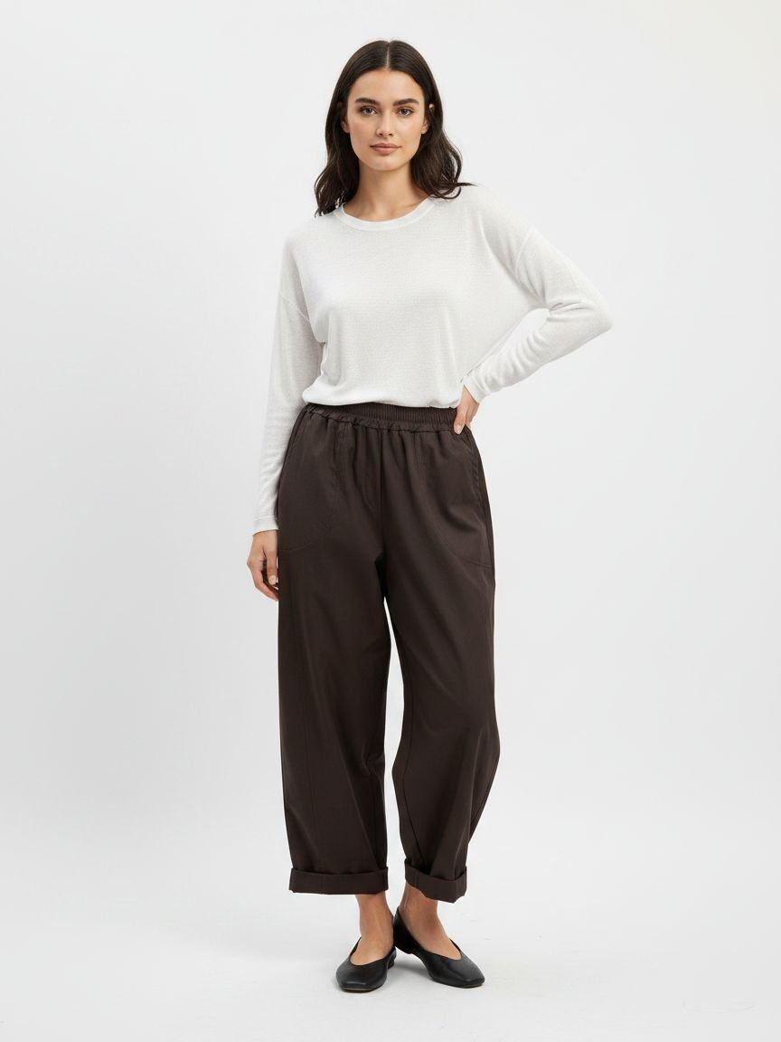 A woman modeling a white long-sleeve top and dark brown wide-leg cropped pants with black flats, standing against a light gray studio background.