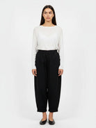 A woman stands against a plain light gray studio background wearing a white long-sleeve top and loose black cropped pants with black flats.