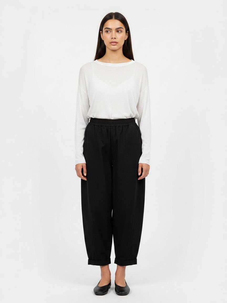 A woman stands against a plain light gray studio background wearing a white long-sleeve top and loose black cropped pants with black flats.