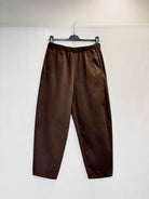 A pair of dark brown wide-leg trousers hanging from a black hanger against a plain white wall.