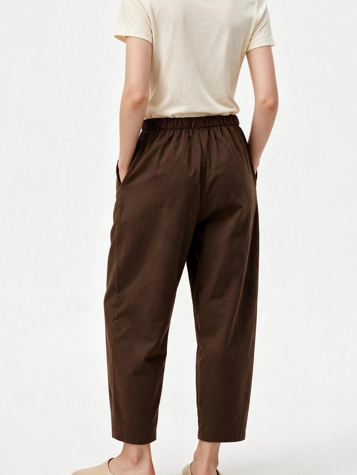 A person shown from the back wearing dark brown cropped pants with an elastic waistband and side pockets, paired with a light cream short-sleeve top and beige slip-on shoes, against a plain white studio background.