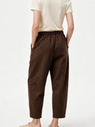 A person shown from the back wearing dark brown cropped pants with an elastic waistband and side pockets, paired with a light cream short-sleeve top and beige slip-on shoes, against a plain white studio background.