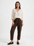 A woman modeling a cream-colored long-sleeve button-up shirt and dark brown cropped trousers with matching brown loafers, set against a plain white studio background.
