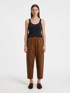 A woman stands against a plain light gray studio background wearing a dark navy sleeveless tank top, brown cropped trousers, and dark brown loafers.