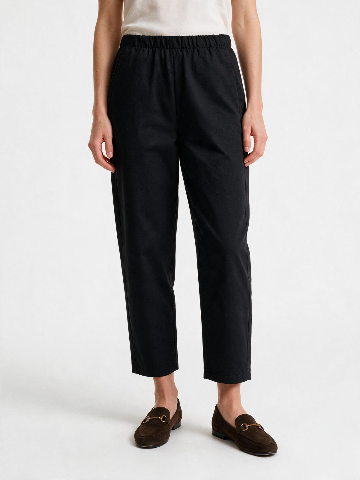 A model is shown from the waist down wearing black cropped trousers with an elastic waistband, paired with brown suede loafers and a cream top, against a plain white studio background.