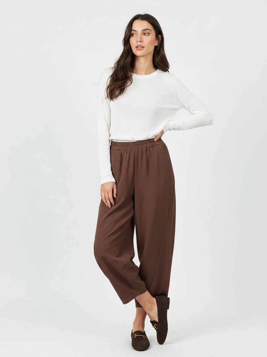 A woman modeling a white long-sleeve knit top and brown wide-leg cropped pants with dark brown loafers, standing against a light gray studio background.