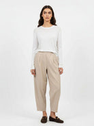 A woman stands against a plain light gray studio background wearing a white long-sleeve top, beige wide-leg cropped pants, and dark brown loafers.