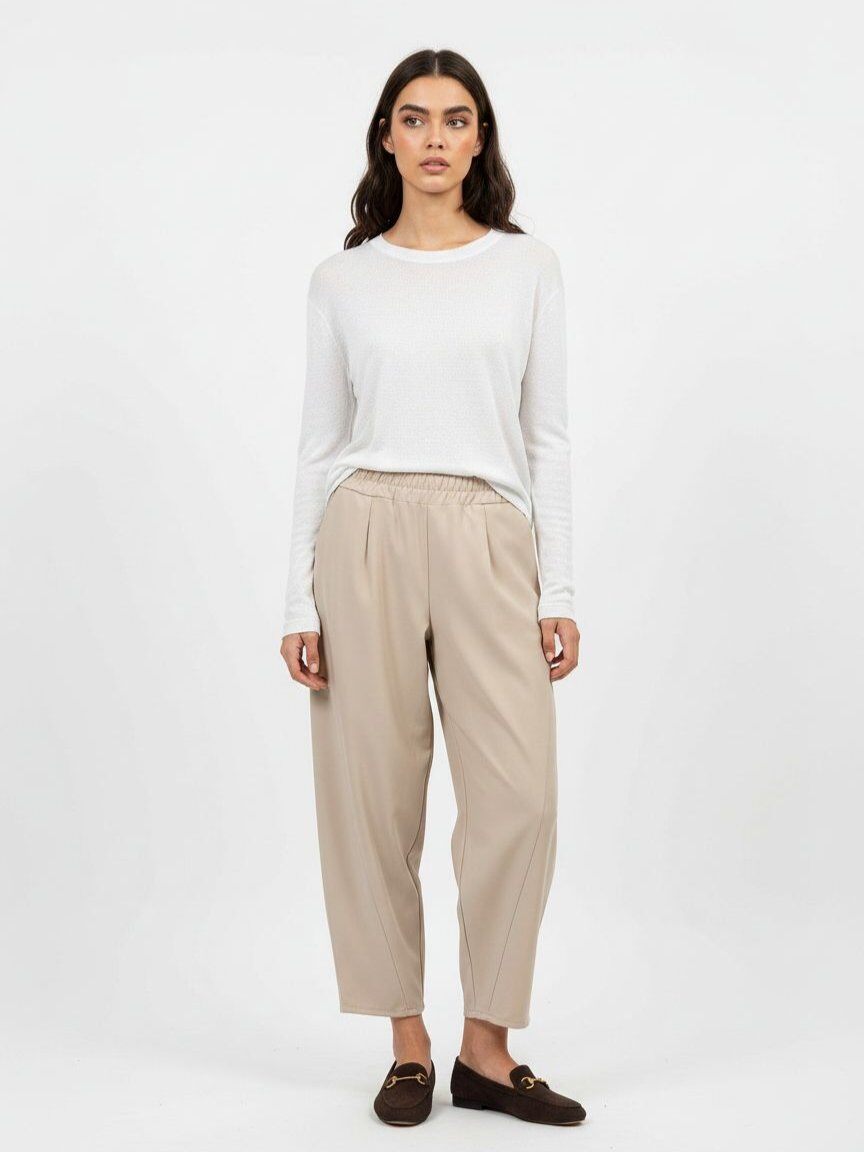 A woman stands against a plain light gray studio background wearing a white long-sleeve top, beige wide-leg cropped pants, and dark brown loafers.