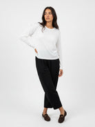 A woman modeling a white long-sleeve crewneck top and black cropped trousers, paired with dark brown loafers, against a plain white studio background.