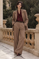A woman stands on an outdoor stone terrace wearing a brown and beige checkered blazer and matching wide-leg trousers, paired with a dark burgundy bow-neck blouse and dark pointed heels.