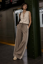 A woman stands in a subway station next to a dark green metal pillar, wearing a sleeveless cream button-up top and wide-leg taupe pants with white side stripes.