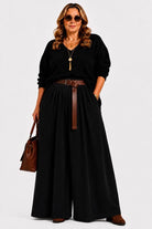 A woman modeling a black long-sleeve top and matching wide-leg pants with a brown belt, sunglasses, a gold pendant necklace, and a brown handbag against a light gray studio background.