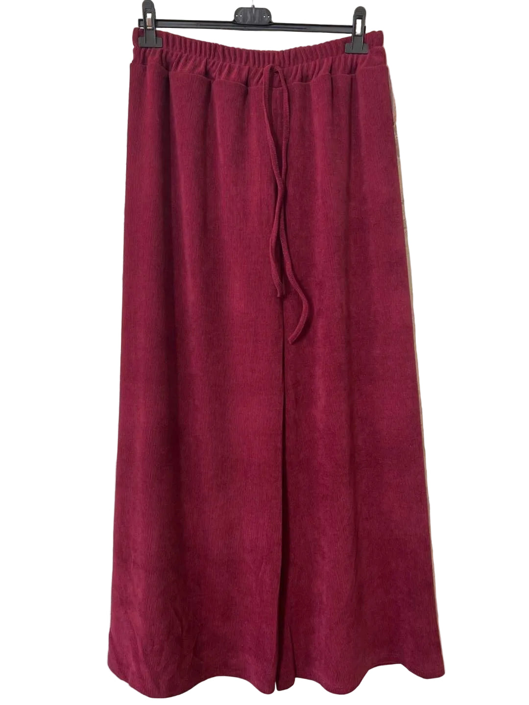 A pair of wide-leg, high-waisted pants in a deep burgundy or maroon corduroy fabric, hanging on a black hanger against a textured gray concrete wall.