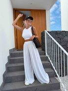 A woman posing on outdoor stone stairs wearing a white sleeveless crop top and matching wide-leg pants, a black cap, sunglasses, and carrying a black shoulder bag.