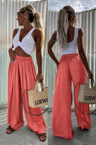A woman shown from the front and back wearing a white twisted-strap crop top and flowing coral wide-leg pants, standing outdoors against a corrugated metal wall and holding a woven Loewe tote bag.