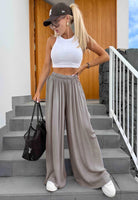 A woman stands on outdoor stone steps wearing a white cropped tank top, loose taupe wide-leg pants, a dark baseball cap, sunglasses, and white sneakers, while holding a black tote bag.