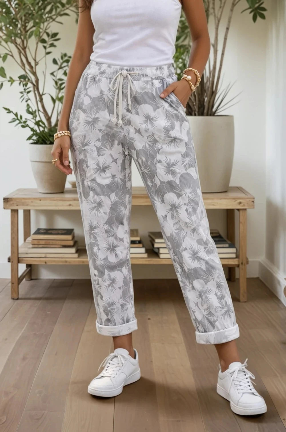 Person wearing gray floral pants and white sneakers in a home setting with plants and books.