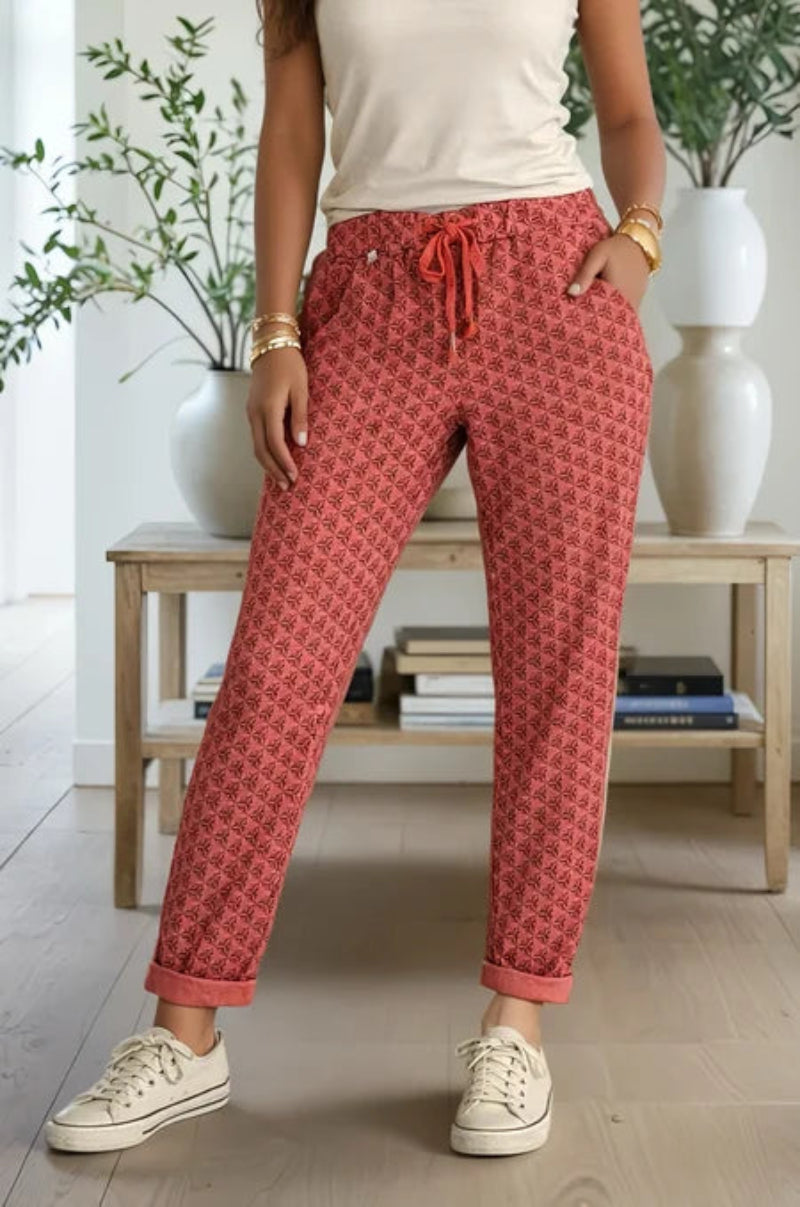 Person wearing coral patterned pants with a white top and white sneakers in a room with plants and books.