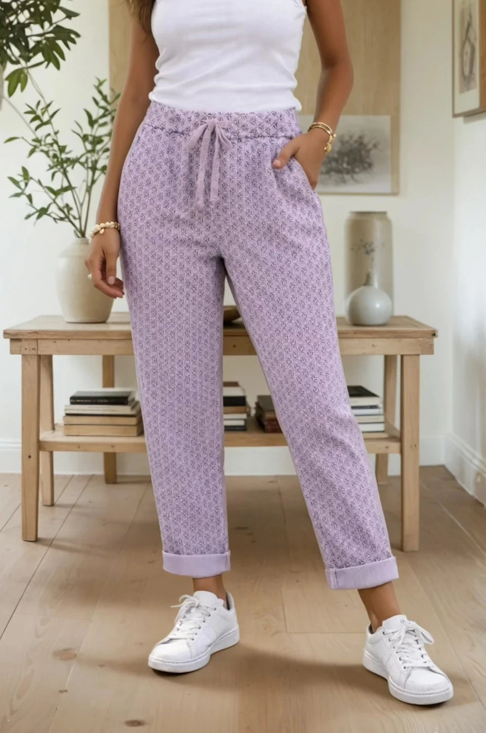 Person wearing lilac patterned pants and white sneakers in a room with wooden furniture and plants.