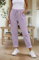 Person wearing lilac patterned pants and white sneakers in a room with wooden furniture and plants.