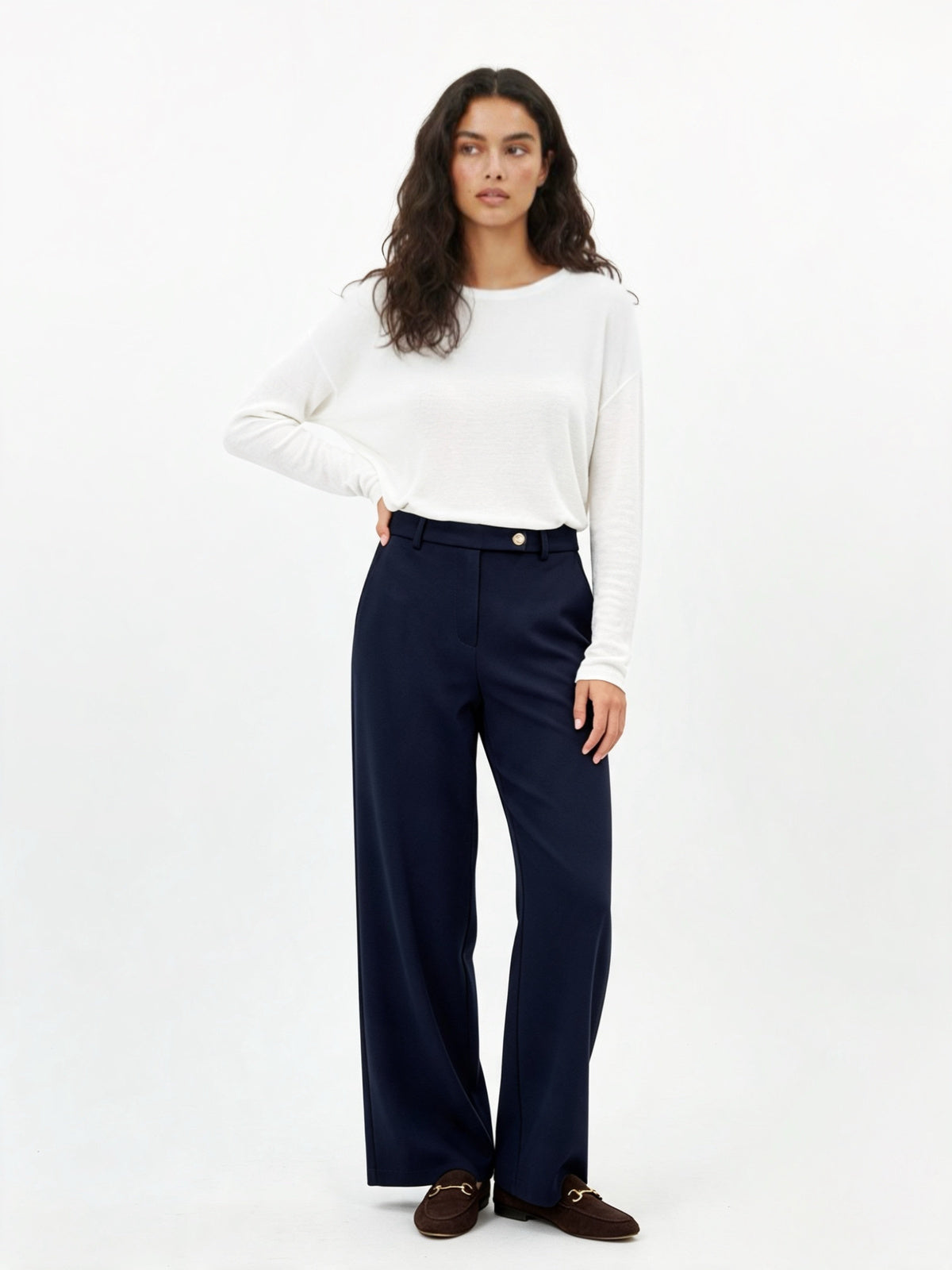 A woman modeling a white long-sleeve crewneck top and navy blue trousers, paired with dark brown loafers, against a plain white studio background.
